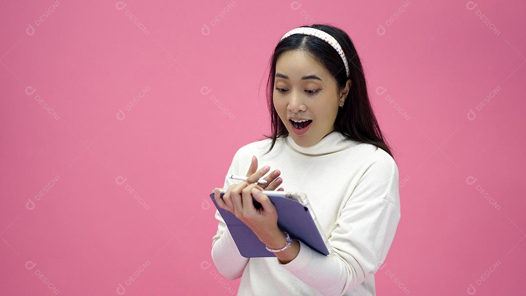 Mulher asiática sorrindo e usando o celular com tablet e animada para fazer compras online em fundo rosa isolado.