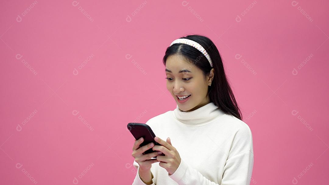 Mulher asiática sorrindo e usando o celular com tablet e animada para fazer compras online em fundo rosa isolado.