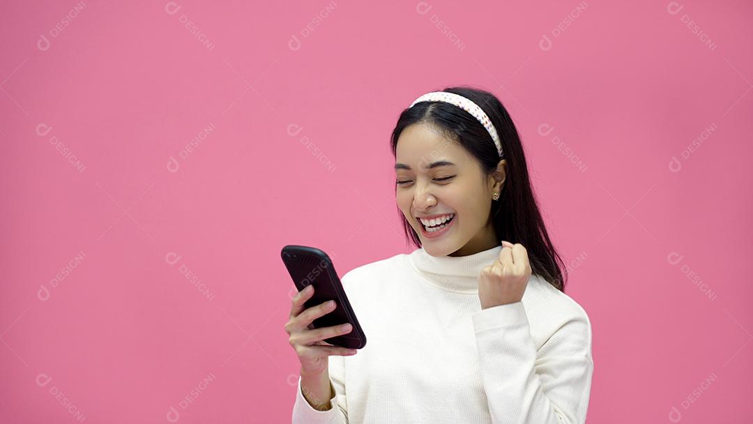 Mulher asiática sorrindo e usando o celular com tablet e animada para fazer compras online em fundo rosa isolado.