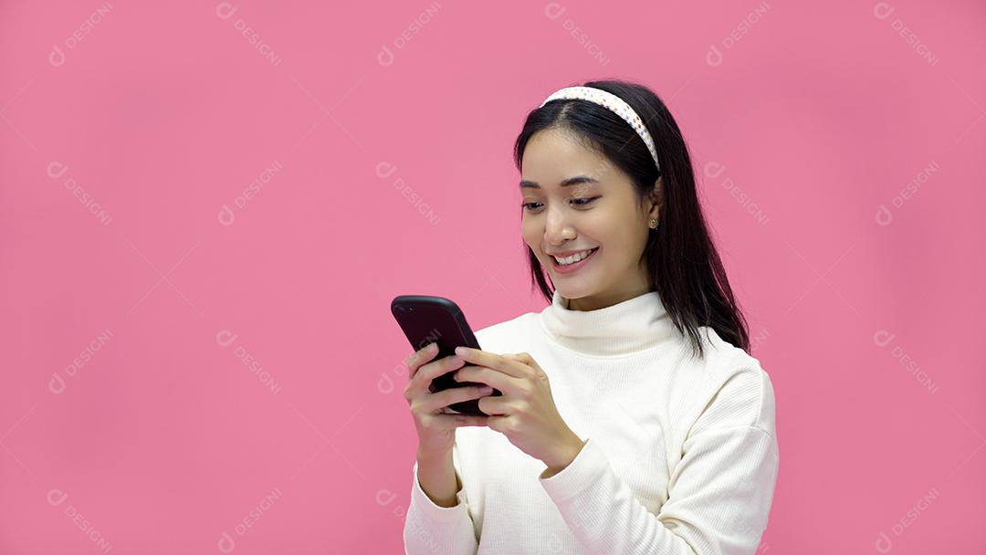 Mulher asiática sorrindo e usando o celular com tablet e animada para fazer compras online em fundo rosa isolado.