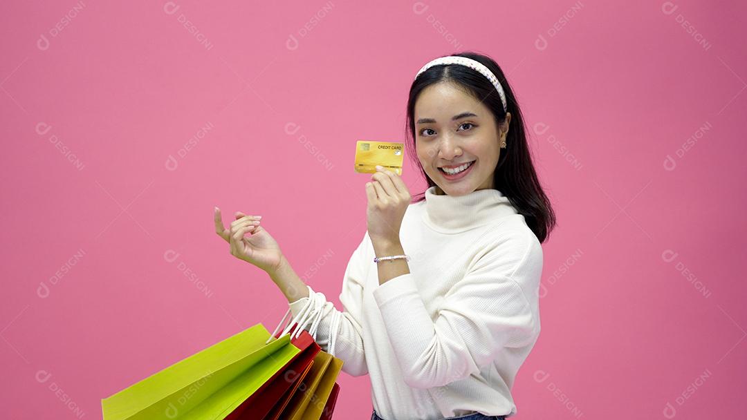 Mulher jovem feliz sorrindo e segurando sacolas de compras e cartão de crédito enquanto fazia algumas compras em um fundo de estúdio rosa