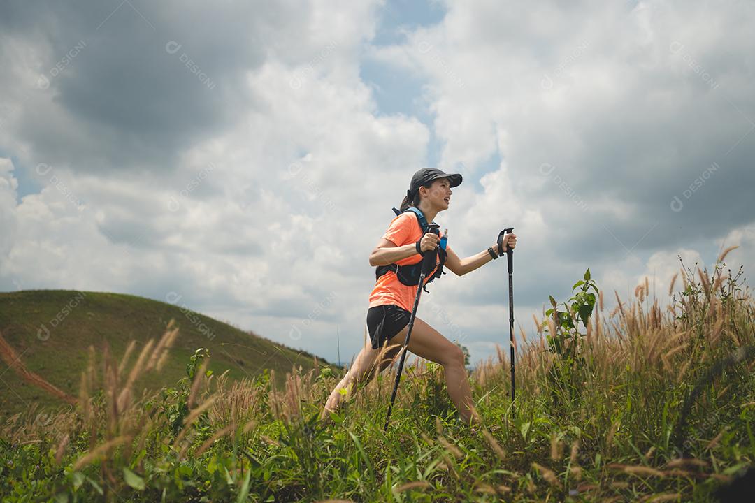 Trilha ativa de mulheres jovens atravessando um prado em uma trilha gramada no alto das montanhas à tarde com vara de trekking