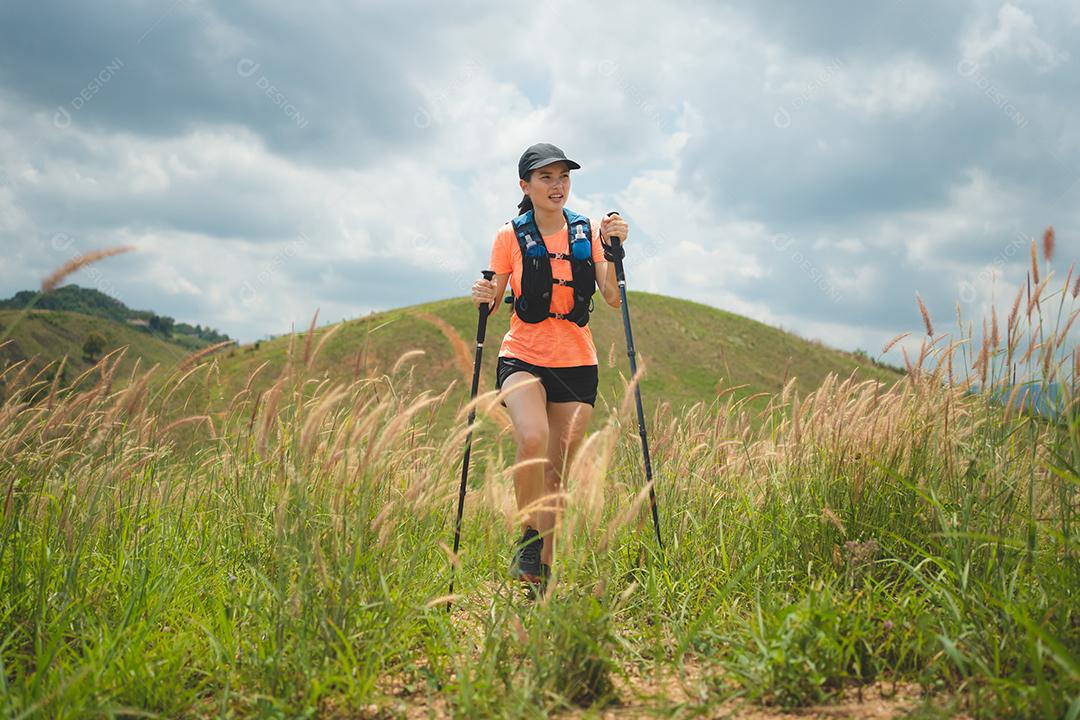 Trilha ativa de mulheres jovens atravessando um prado em uma trilha gramada no alto das montanhas à tarde com vara de trekking
