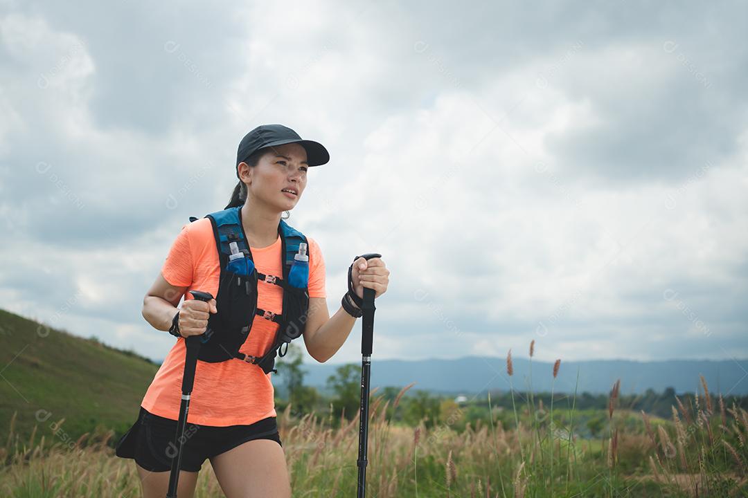 Trilha ativa de mulheres jovens atravessando um prado em uma trilha gramada no alto das montanhas à tarde com vara de trekking