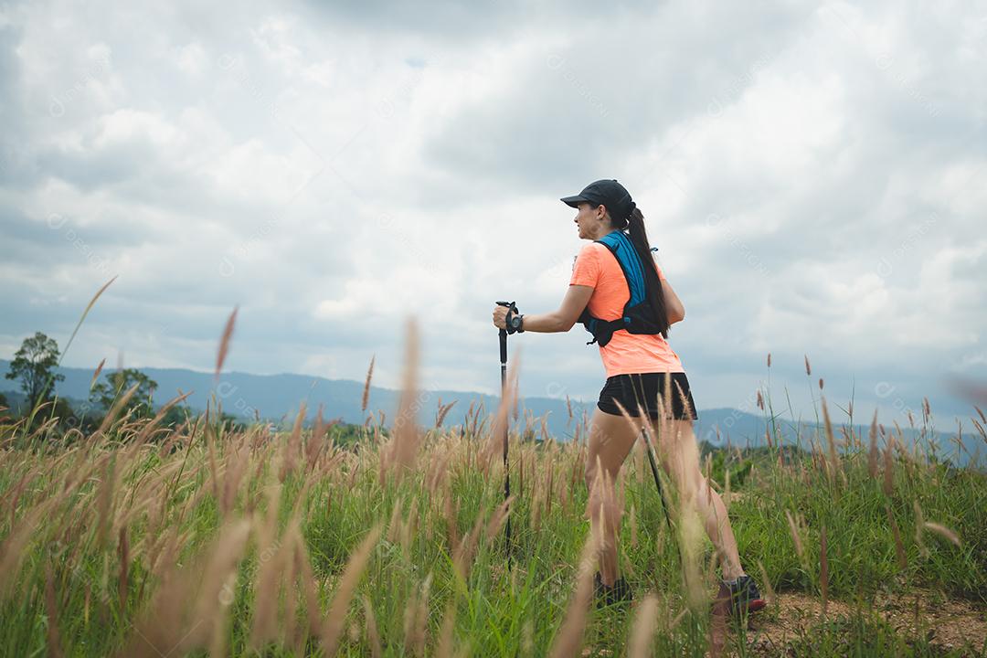 Trilha ativa de mulheres jovens atravessando um prado em uma trilha gramada no alto das montanhas à tarde com vara de trekking