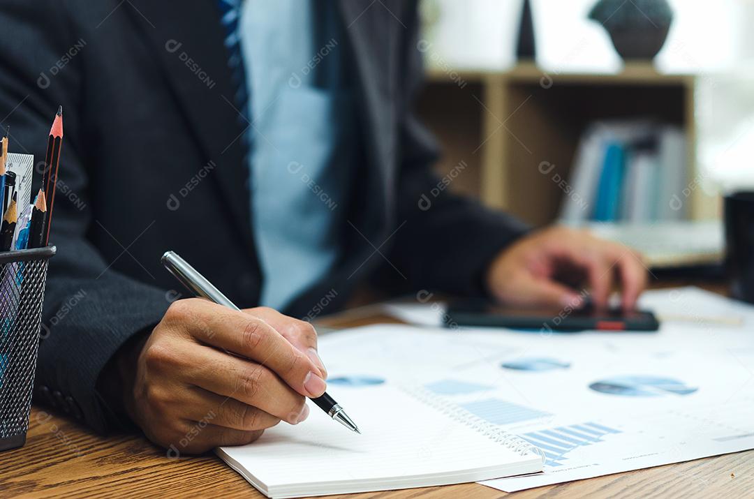 Empresário segurando caneta escrevendo um livro e gráfico de documento contabilidade finanças papel análise gráfico investimento de marketing e economia de relatório na mesa no escritório.