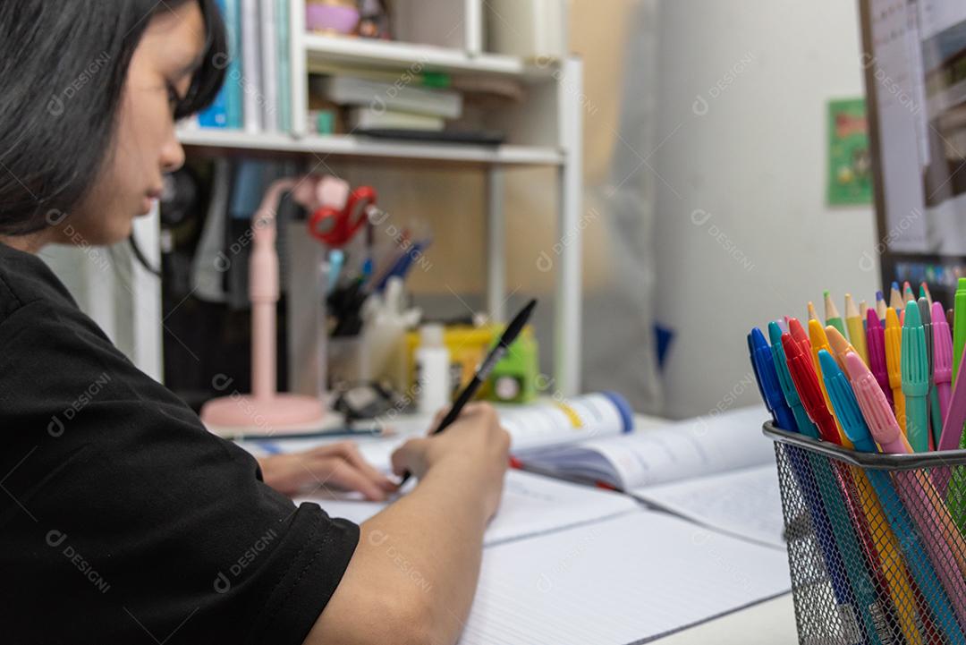 Aluna asiática está escrevendo lição de casa e lendo livro na mesa