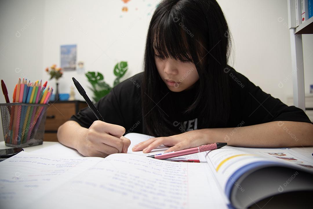 Aluna asiática está escrevendo lição de casa e lendo livro na mesa