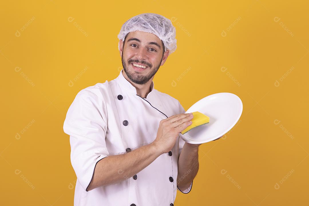 homem chef de cozinha segurando uma esponja de lavar louça e um prato sob fundo amarelo