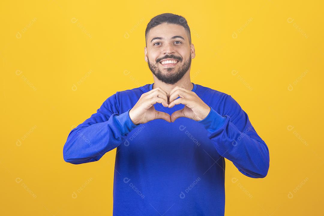 Retrato de jovem sorridente mantém as mãos no peito em sinal de forma de coração, expressa simpatia.