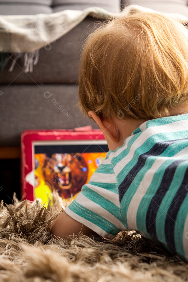 Criança assiste vídeos em um tablet, deitada no tapete da sala, brincando, assistindo e aprendendo