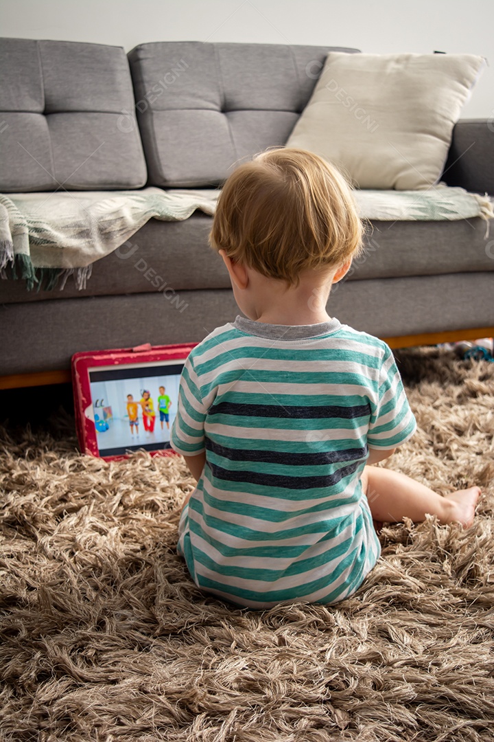 Criança assiste a vídeos em um tablet, sentada no tapete da sala, brincando, assistindo e aprendendo