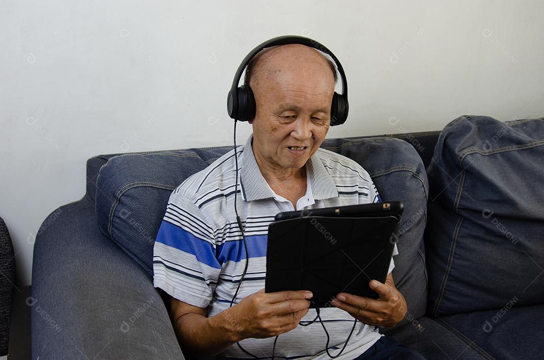 Homem idoso segurando um tablet no sofá ouvindo música com fones de ouvido e mídias sociais.