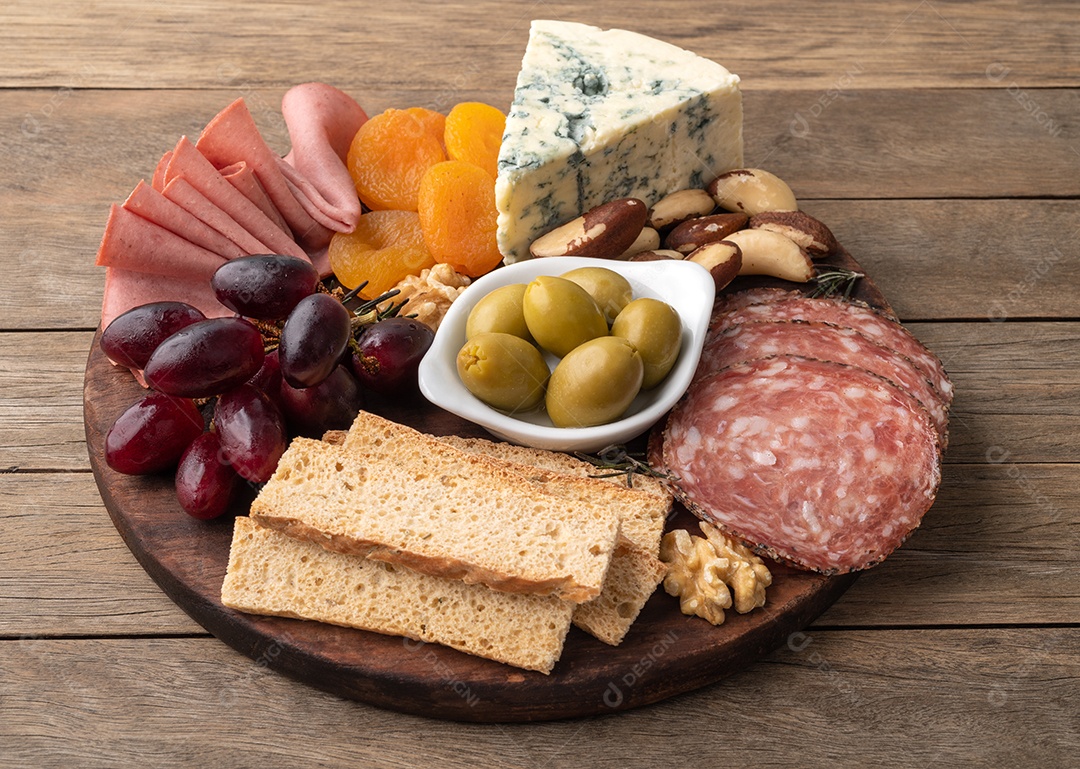 Tábua de aperitivos com queijo, nozes, frutas, torradas e charcutaria sobre a mesa de madeira.