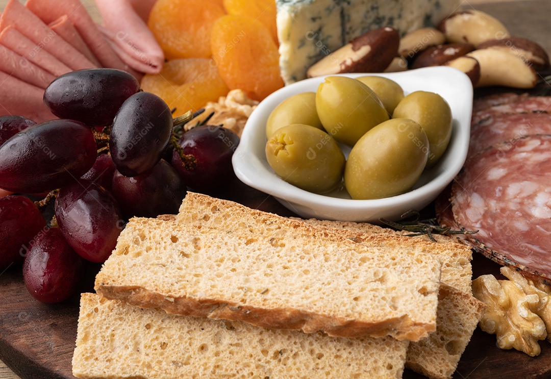 Tábua de aperitivos com queijo, nozes, frutas, torradas e charcutaria sobre a mesa de madeira.