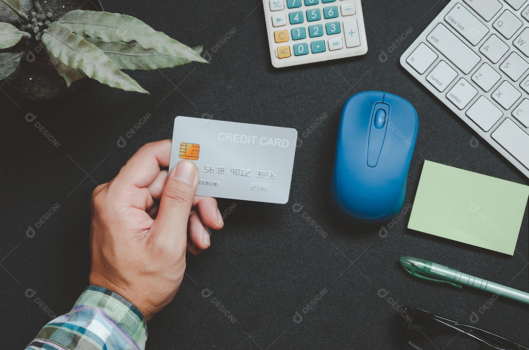 Homem de vista superior segurando um cartão de crédito e na mesa. Bloco de notas e caneta do computador portátil Negócios de compras online