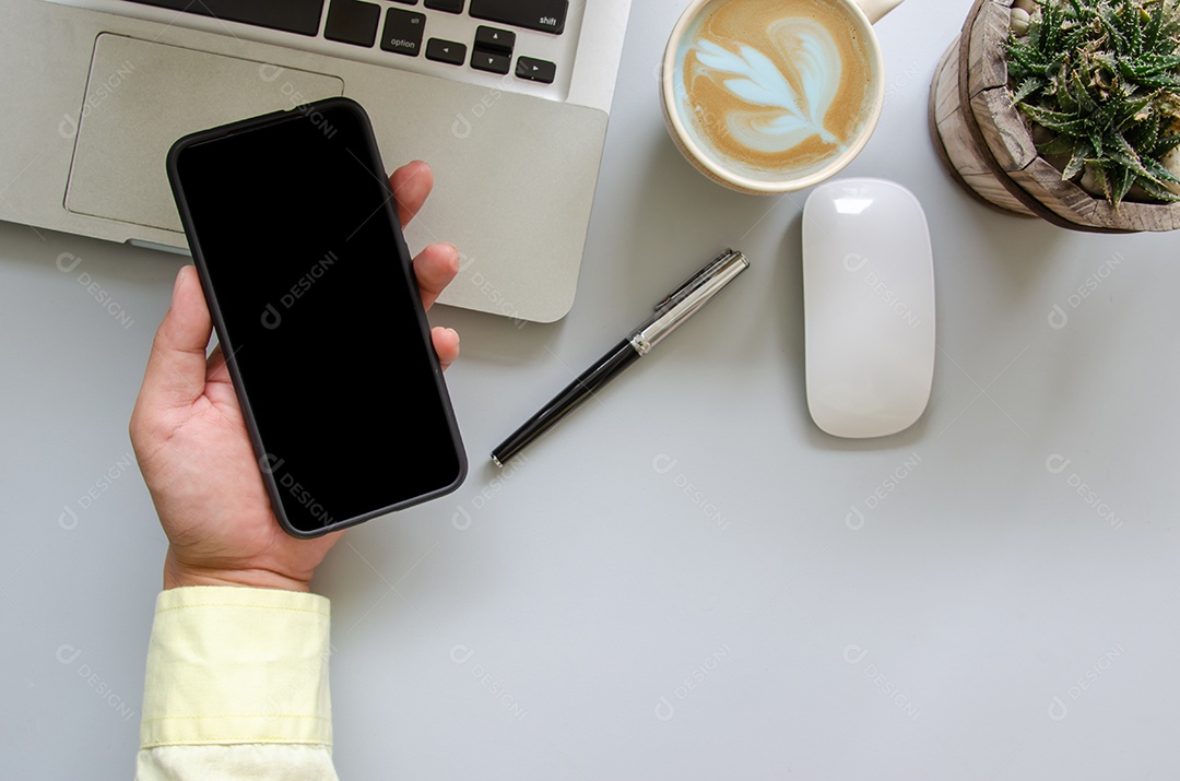 Male hand holding smartphone, computer, pen, coffee, mouse on office desk with copy space