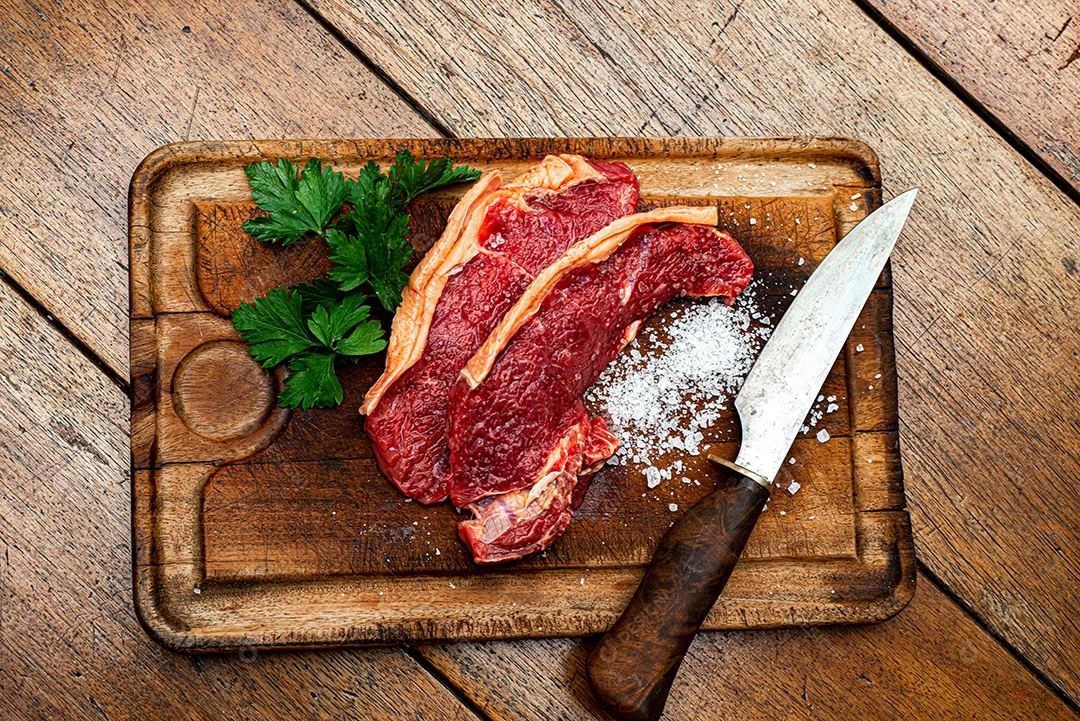 Bife de picanha de carne fresca crua, corte tradicional brasileiro com salsa, sal grosso e faca na placa de madeira. Visão do topo