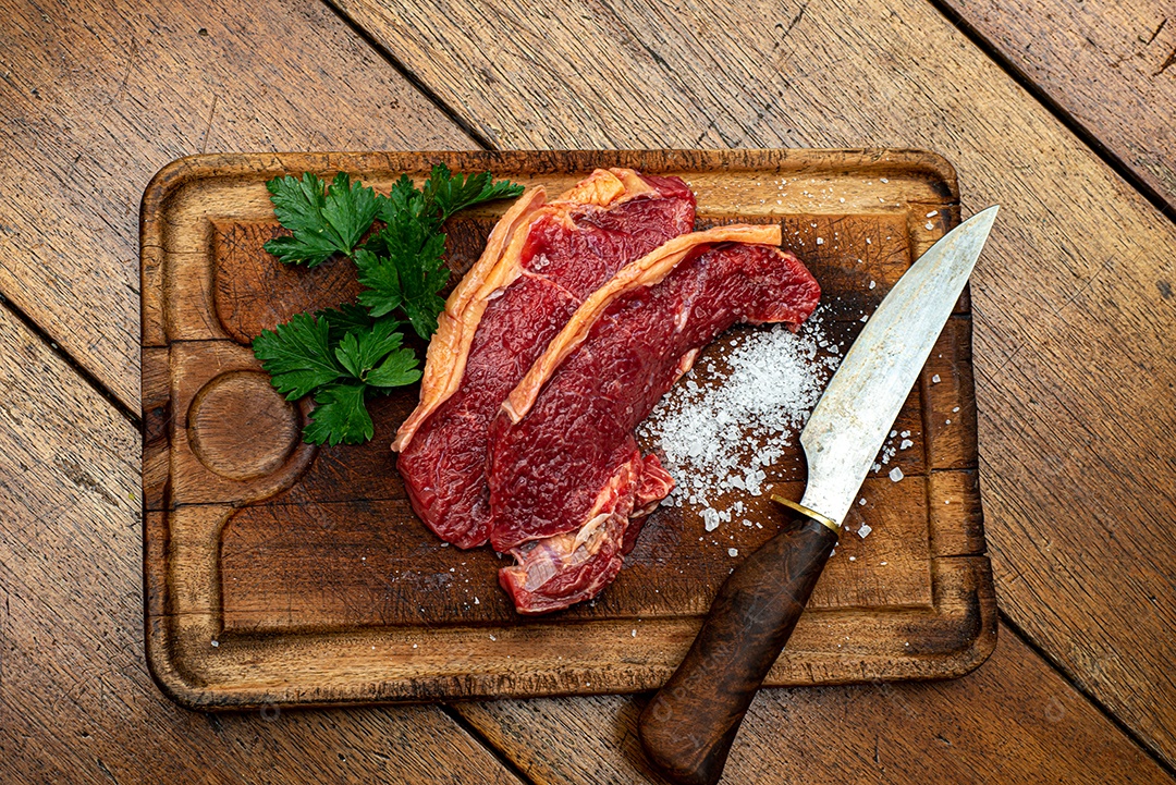 Bife de picanha de carne fresca crua, corte tradicional brasileiro com salsa, sal grosso e faca na placa de madeira. Visão do topo