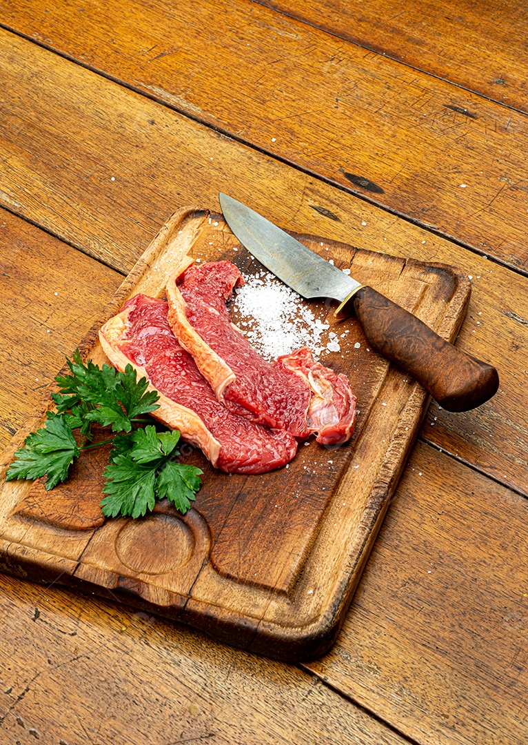 Bife de picanha de carne fresca crua, corte tradicional brasileiro com salsa, sal grosso e faca na placa de madeira. Visão do topo