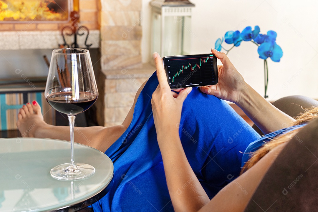 Jovem investidora do mercado de ações segurando um gráfico de crescimento do índice do aplicativo de investimento em telefone - Mulher de negócios do milênio investe na bolsa de valores sentada na sala de estar bebendo um vinho - conceito de finanças