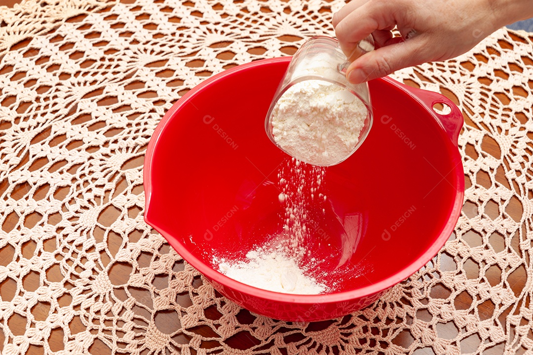 Mão colocando açúcar com colher em tigela de plástico vermelha com farinha de trigo. Fundo de toalha de mesa de crochê. Vista do topo. Copie o espaço.