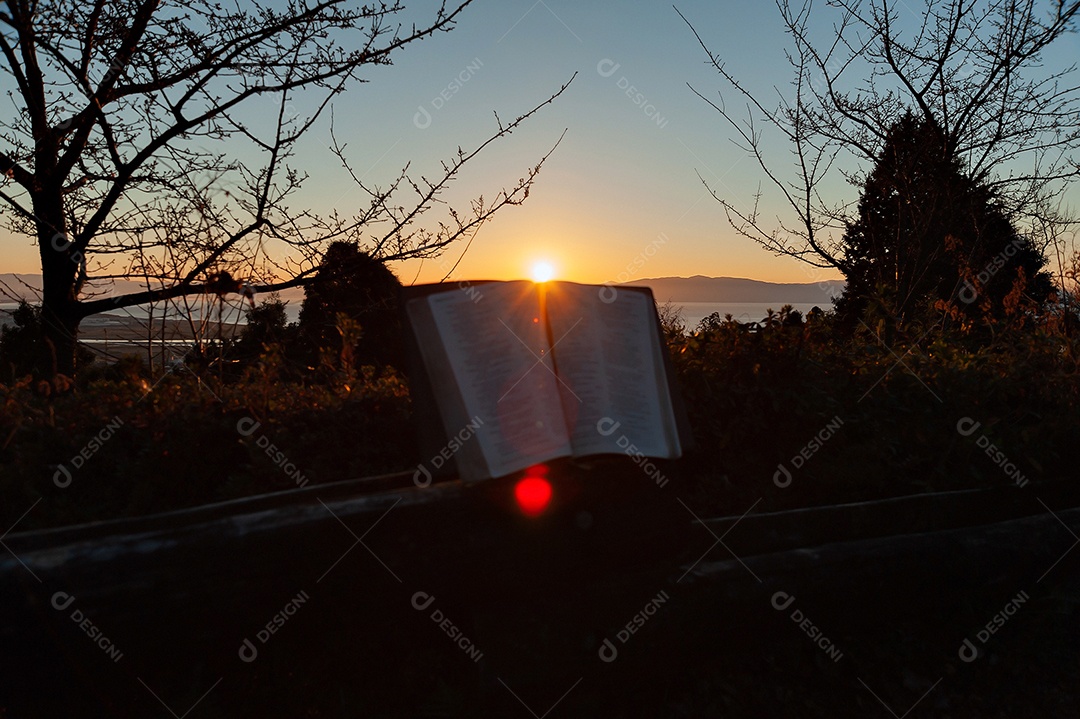 Bíblia Sagrada aberta ao amanhecer com luz solar e reflexo de lente. Tiro horizontal.