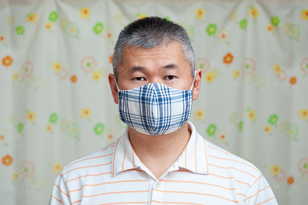 Vista frontal do homem asiático de meia idade sentado no sofá usando máscara facial de tecido DIY para proteção contra coronavírus (COVID-19) e outras doenças infecciosas. Fundo de cortina floral.