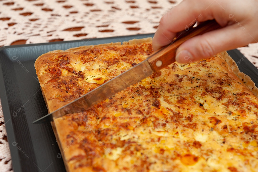 Vista lateral da mão de mulher cortando torta salgada de liquidificador brasileiro - comida tradicional brasileira feita com atum, ovo cozido, milho, frango desfiado, legumes etc. Fundo de toalha de crochê artesanal.