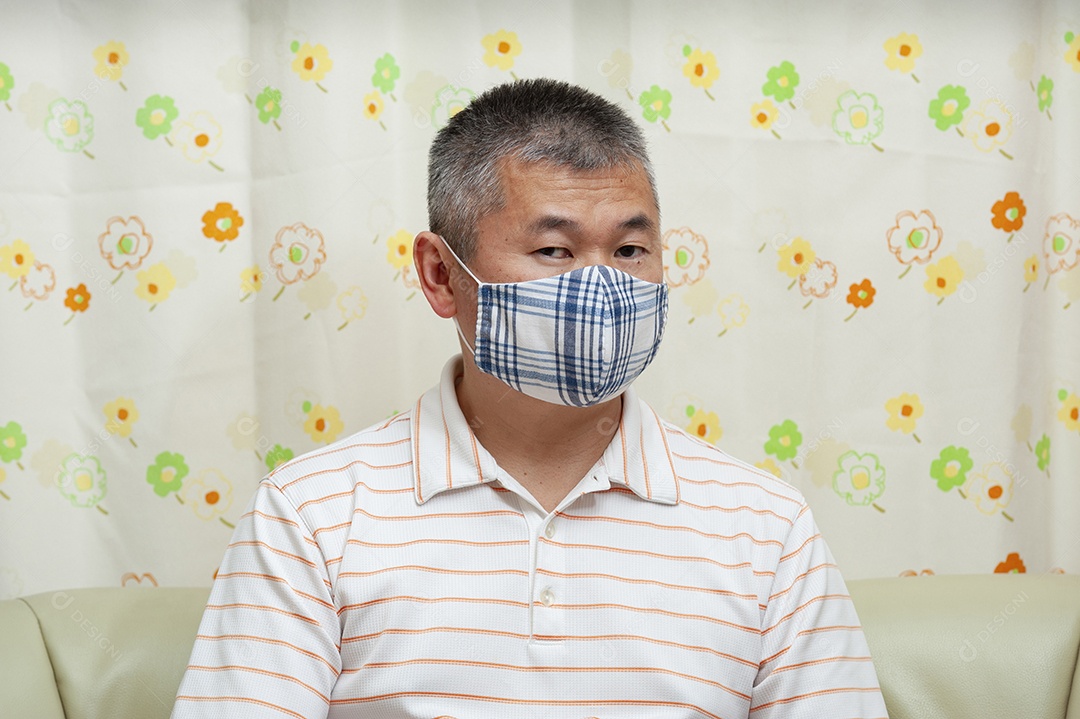 Vista frontal do homem asiático de meia idade sentado no sofá usando máscara facial de tecido DIY para proteção contra coronavírus (COVID-19) e outras doenças infecciosas. Fundo de cortina floral.