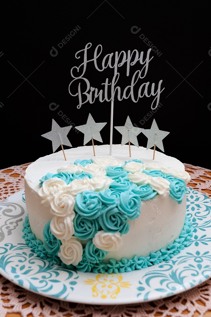 Bolo de aniversário coberto com glacê branco e azul. Decorado com formas de estrelas e a frase: Feliz Aniversário. Isolado em fundo preto.