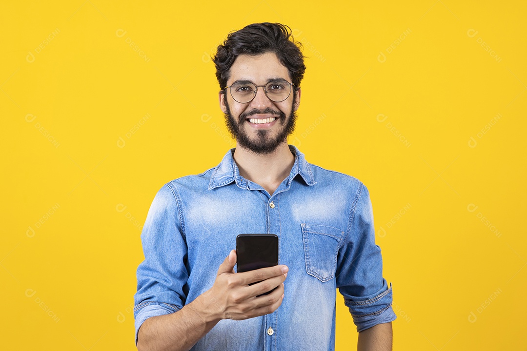 Homem jovem segurando celular barbudo sobre fundo amarelo