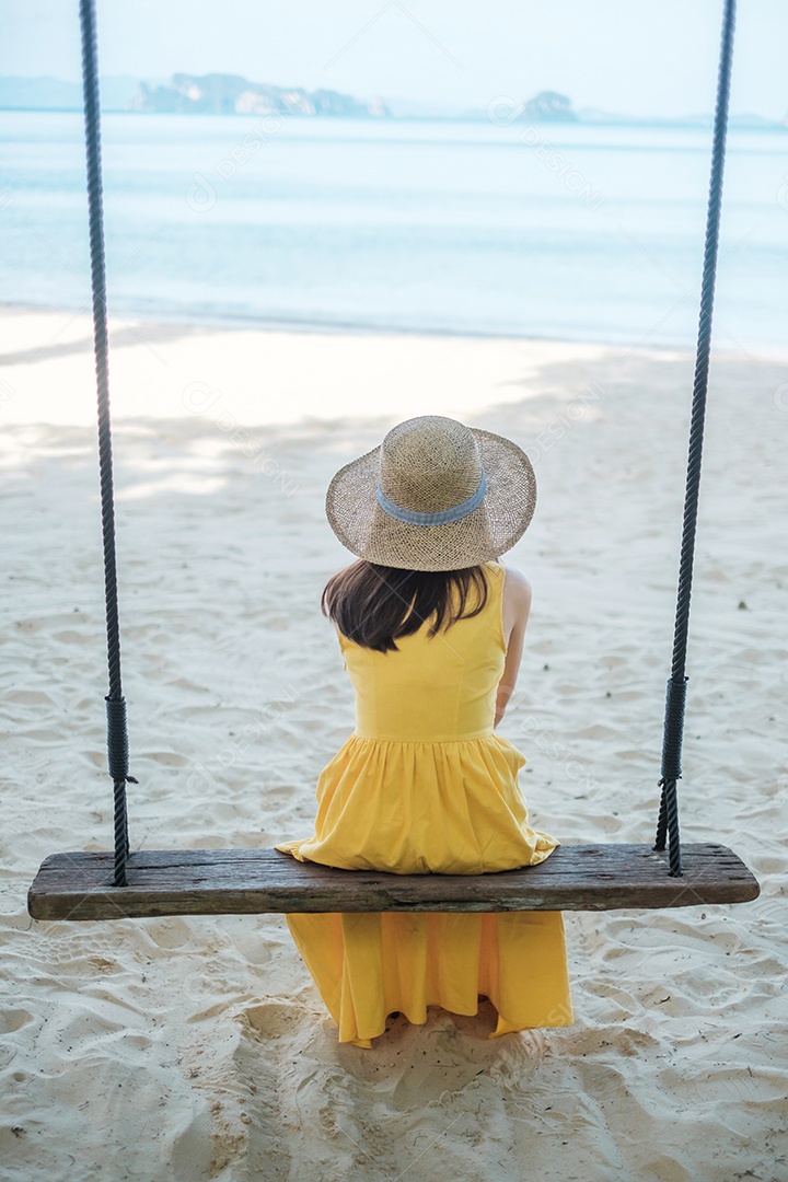 Happy Woman Traveler está relaxando em um balanço de madeira na praia paradisíaca. Turista feminina em vestido amarelo