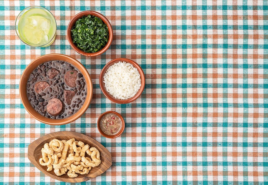 Feijoada típica brasileira com arroz, couve, farofa, caipirinha e torresmo sobre tecido xadrez com espaço para cópia.