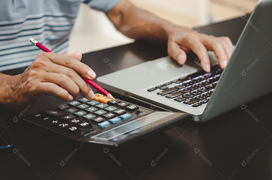 Mão segurando um lápis, calculadora e teclado de computador na mesa.