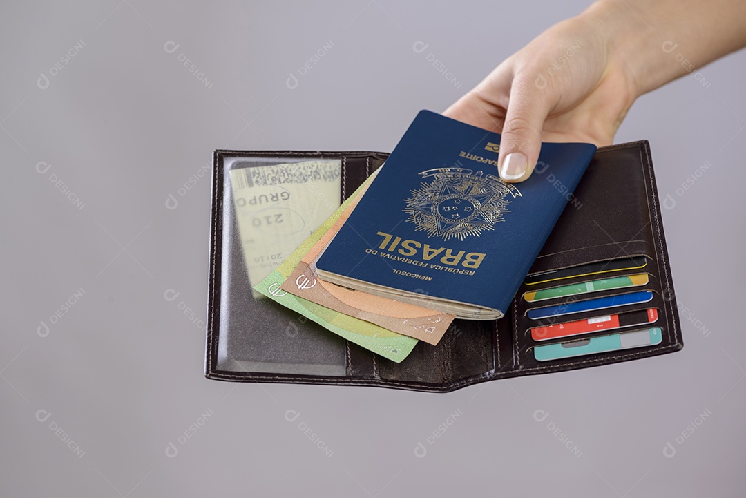Passaporte brasileiro em uma mão com carteira, cartões de crédito e notas de euro em um fundo branco.