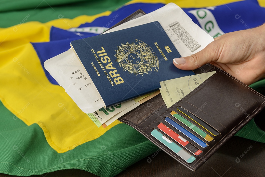 Mão segurando o passaporte brasileiro sobre a mesa com bandeira brasileira ao fundo.