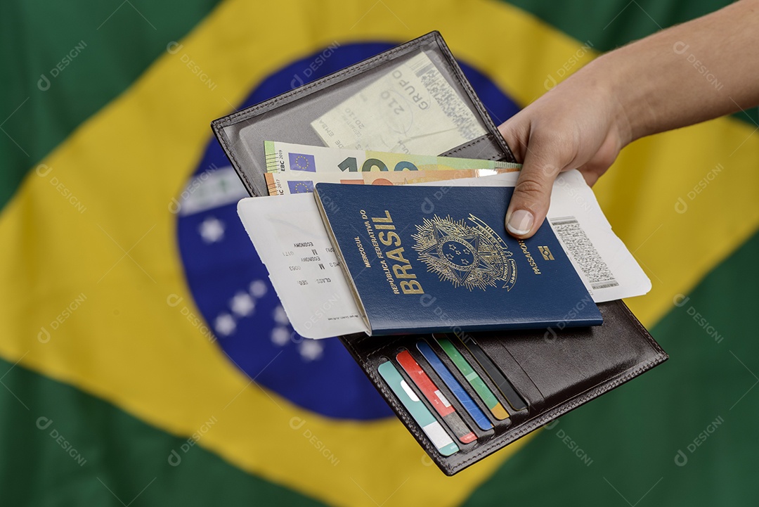 Mão segurando passaporte brasileiro com bandeira brasileira no fundo desfocado.
