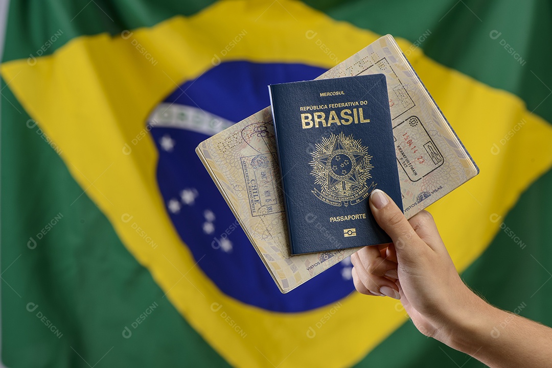 Mão segurando passaporte brasileiro com bandeira brasileira no fundo desfocado.
