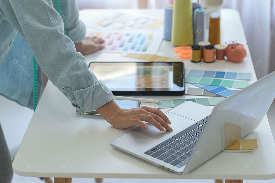 Designer de roupas, alfaiate projetando com laptops e tablets, suprimentos de costura na mesa da oficina.