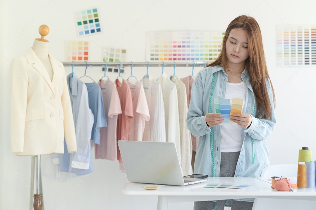 Jovem costura no espaço de trabalho de um designer com equipamento de design, laptop e smartphone em uma oficina.