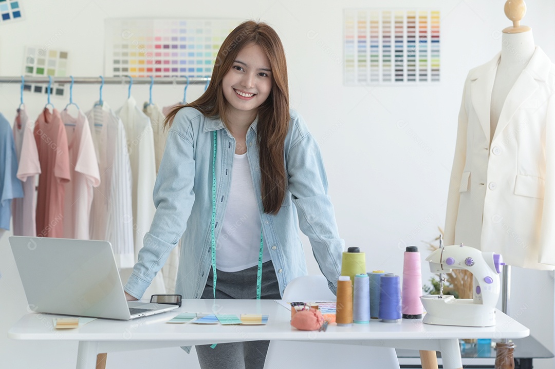 Jovem costura no espaço de trabalho de um designer com equipamento de design, laptop e smartphone em uma oficina.
