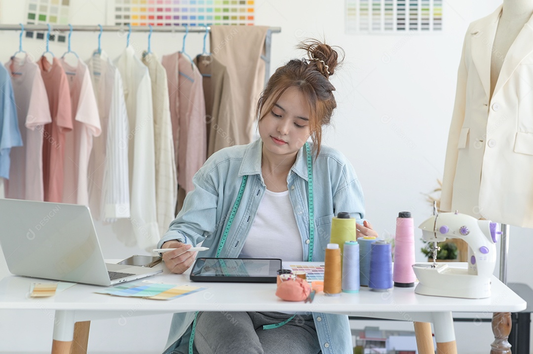 Jovem costura no espaço de trabalho de um designer com equipamento de design, laptop e smartphone em uma oficina.