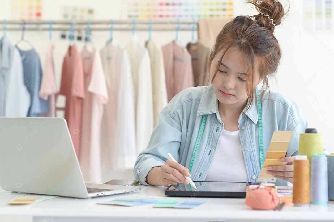 Jovem costura no espaço de trabalho de um designer com equipamento de design, laptop e smartphone em uma oficina.