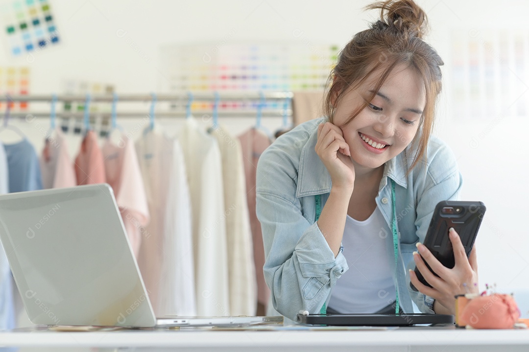 Jovem costura no espaço de trabalho de um designer com equipamento de design, laptop e smartphone em uma oficina.