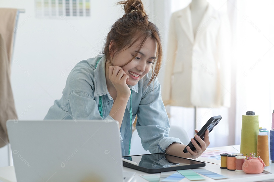 Jovem costura no espaço de trabalho de um designer com equipamento de design, laptop e smartphone em uma oficina.