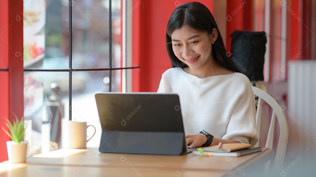 Jovem usando um laptop para videoconferência em um café, filmado do lado de fora através do vidro.