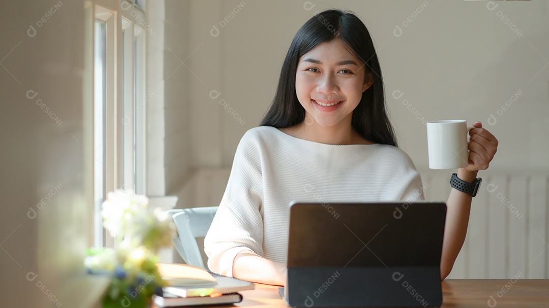 Moça segura uma xícara de café e usa um laptop. Ela olhou para a câmera e sorriu feliz.