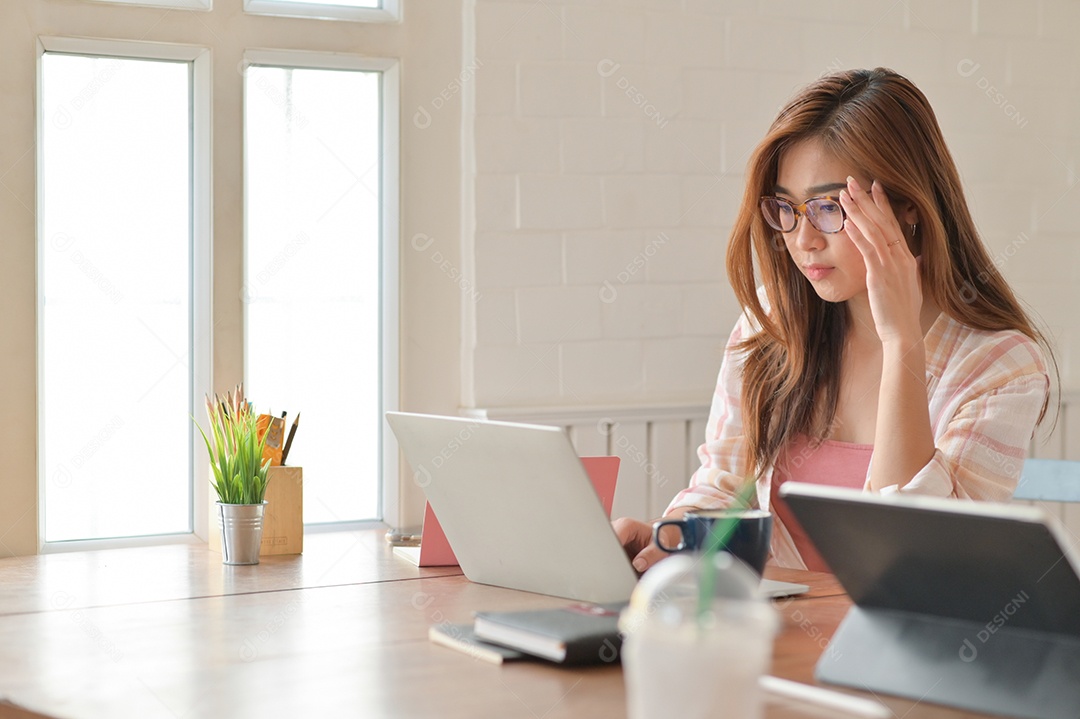 Estudante asiática, segurando os óculos com cuidado enquanto olha para o laptop para se preparar para estudos de pós-graduação.