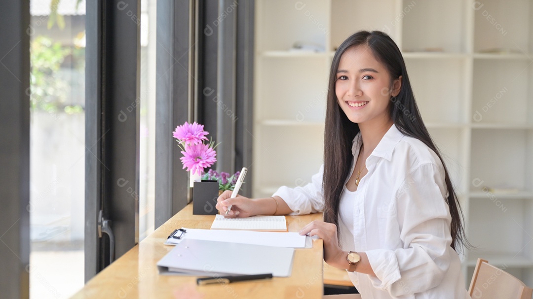 retrato de uma mulher asiática usando um vestido branco segurando uma caneta para escrever notas em seu caderno em um escritório confortável. Ela olhou para a câmera e sorriu.
